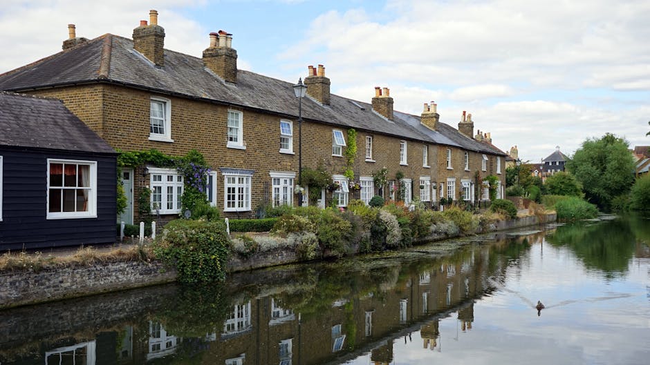 A row of traditional terraced houses constructed with yellow brick, with pitched slate roofs and multiple chimneys, situated along the edge of a calm river or canal in West Green. The front gardens feature well-maintained greenery, small shrubs, and flowering plants, some supported by white wooden fences. Several multi-pane sash windows and white doors are visible, with climbing plants and decorative features around the entrances. The reflection of these houses is seen clearly in the water. To the left, there is a small dark-colored wooden shed with a window, positioned at the corner of the property. The scene is illuminated by natural daylight, with a partly cloudy sky overhead. This image captures the typical residential architecture and picturesque setting seen in West Green, relevant for house removals, packing, and moving logistics companies like Man with Van West Green, preparing for a home relocation or furniture transport process.