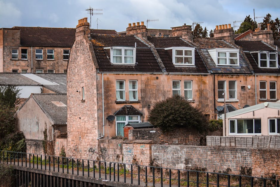A row of traditional terraced houses constructed with yellow brick, with pitched slate roofs and multiple chimneys, situated along the edge of a calm river or canal in West Green. The front gardens feature well-maintained greenery, small shrubs, and flowering plants, some supported by white wooden fences. Several multi-pane sash windows and white doors are visible, with climbing plants and decorative features around the entrances. The reflection of these houses is seen clearly in the water. To the left, there is a small dark-colored wooden shed with a window, positioned at the corner of the property. The scene is illuminated by natural daylight, with a partly cloudy sky overhead. This image captures the typical residential architecture and picturesque setting seen in West Green, relevant for house removals, packing, and moving logistics companies like Man with Van West Green, preparing for a home relocation or furniture transport process.