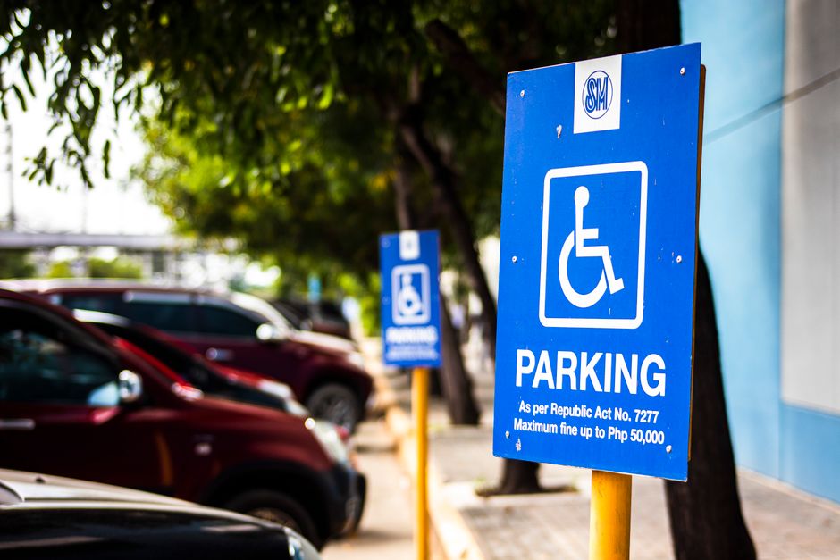 A clear view of a blue parking sign with a white wheelchair symbol indicating accessible parking, situated on a yellow pole beside a row of parked vehicles on a street. The sign includes information about parking regulations and fines, referencing the Republic Act No. 7277, which applies to West Green. Behind the sign, several cars are parked close together along the pavement, with some partially visible and in various colours including red and black. The background shows blurred green foliage from nearby trees and part of a building with a grey wall. The scene is well-lit with natural daylight, emphasizing the parking signage and the surrounding parked vehicles, relevant to home relocation and furniture transport logistics managed by Man with Van West Green as part of their removals and moving services.