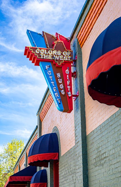 A neon sign attached to the side of a building displays the words 'Colors of the West' in multiple colors, with additional text indicating 'Fountains', 'Gallery', and 'Lighting'. The sign is mounted near the corner of the brick facade, which features two blue and red striped fabric awnings covering windows or entryways. The sign is positioned above the red brickwork and light-colored structural elements, set against a bright blue sky with some green tree foliage visible in the background. The scene reflects urban commercial architecture, potentially related to retail or entertainment venues, and provides a neutral context suitable for illustrating house removals or moving logistics connected to businesses or storefronts, with [COMPANY_NAME] offering similar services.