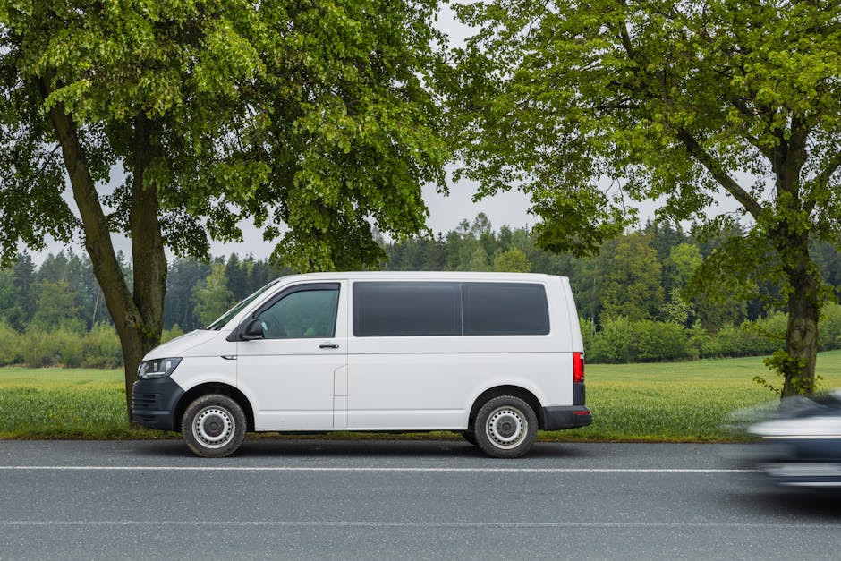 A white panel van parked on a grassy area outside a residential building with brick walls and windows. The van is positioned near a tree with green foliage casting shadows on its side. Part of the rea