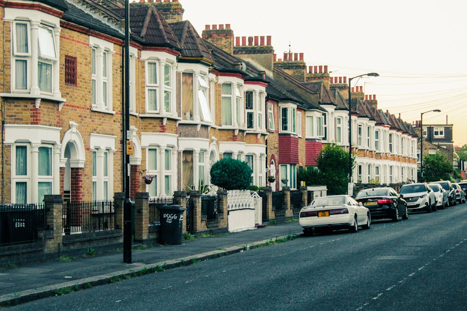 A row of terraced houses in West Green with brick facades, white window frames, and small front gardens enclosed by low wall fences. Several houses have bay windows, some with open window panes, and decorative architectural details around the windows and doors. The pavement along the street is lined with parked cars, including sedans and hatchbacks of various colours. In the foreground, a black wheelie bin with white lettering is visible on the sidewalk. Street lamps and overhead electrical wires run along the road, which appears calm and residential. The lighting suggests it is either early morning or late afternoon. This setting illustrates a typical urban residential area in Haringey, suitable for home relocation or furniture transport services with considerations for parking permits from Haringey Council for moving vans, such as those provided by Man with Van West Green, to facilitate efficient move planning and logistical support during packing and loading processes.