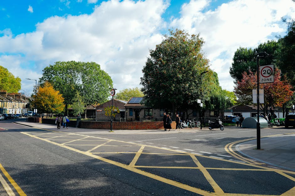 A street view in West Green with a small green park area surrounded by trees with green, yellow, and red foliage, and several pedestrians walking along the pavement near the park. A brick wall surrounds the park, and there are parked bicycles and motorcycles along the sidewalk. In the background, residential buildings with pitched roofs and a variety of parked cars are visible. The sky is partly cloudy with patches of blue. A street sign indicates a 20 mph speed zone. The scene is well-lit with natural daylight, showing the typical environment for vehicle loading and home relocation activities in the West Green area, supported by professional removals services like those provided by Man with Van West Green.