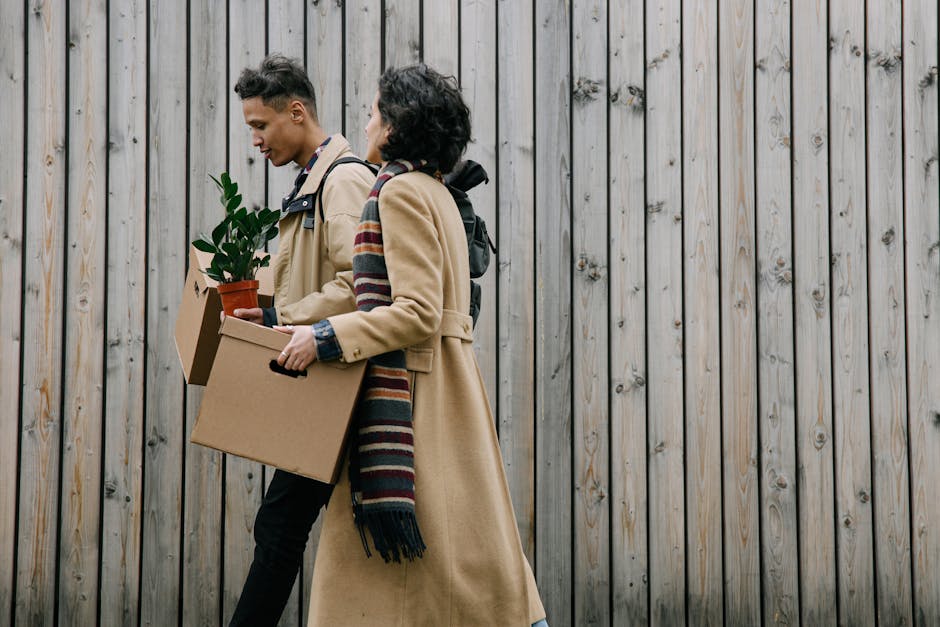 Two individuals, a man and a woman, are walking past a tall wooden fence while carrying boxes and a potted plant, likely during a house move or relocation process. The man is holding a cardboad box with a small potted plant and a larger cardboard box, while the woman is carrying a cardboard box and has a blanket scarf draped over her shoulders. Both are dressed in casual clothing suitable for moving, with the man wearing a beige coat and the woman in a cream-colored coat. A backpack is visible on the woman's back. The scene is outdoors, in front of a vertical wooden fence with natural wood tones, suggesting they are either arriving at or leaving a property as part of a home relocation service. This visual depicts the packing and moving activities typical of professional removals, such as those offered by Man with Van West Green, as part of a comprehensive furniture transport and logistics process.