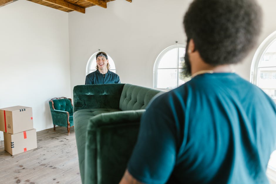 A woman with curly dark hair, dressed in a white sweater and blue jeans, is carrying a large wooden plant pot with green foliage in her right hand, while holding a cardboard box with potted plants and a cardboard box marked 'BOOKS' in her left hand. She is standing at the bottom of a staircase inside a home, near the doorway to an upper room. A person wearing dark clothing and black footwear is partially visible, holding a similar box, indicating a house move or packing process. The staircase has wooden banisters and carpeted steps, with a bicycle visible in the background behind the woman. This scene captures a home relocation activity, part of a packing and moving service, with visible packing materials and multiple items being prepared for transport, typical of a professional removals process by Man with Van West Green.