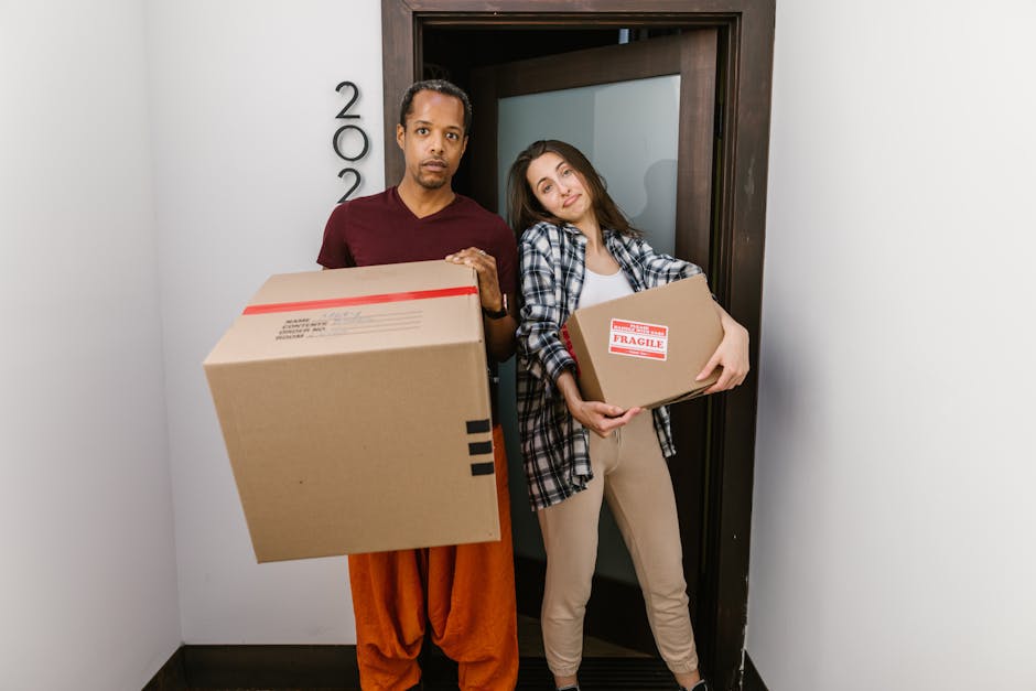 A man and a woman stand side by side in a narrow hallway of a property, each holding a cardboard box; the man holds a large, plain cardboard box with black tape on the edges, while the woman carries a