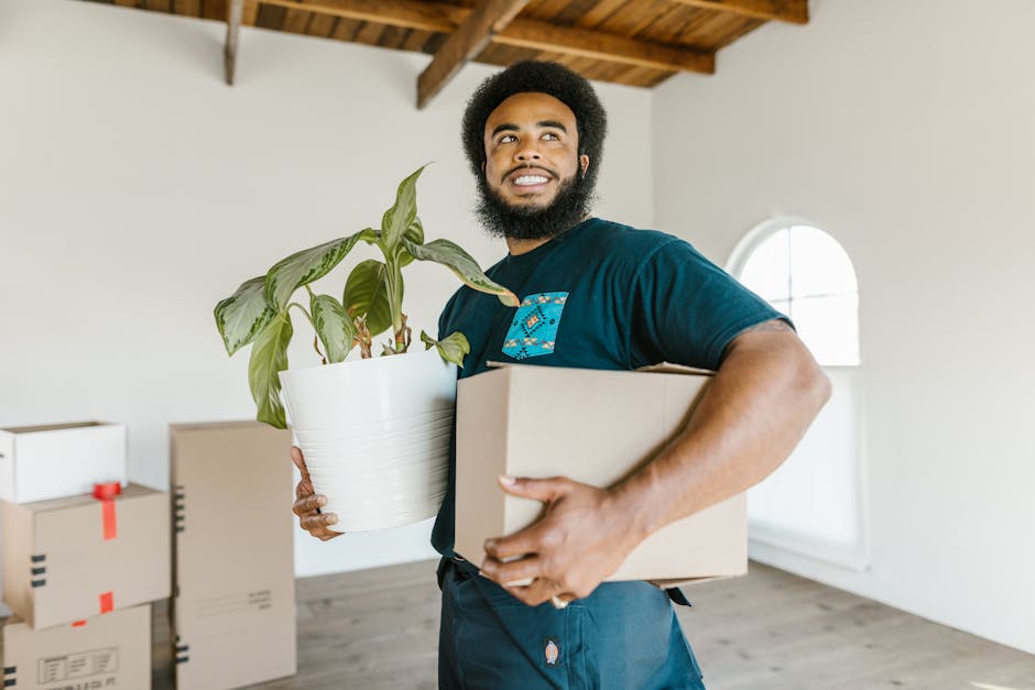 A man and a woman stand side by side in a narrow hallway of a property, each holding a cardboard box; the man holds a large, plain cardboard box with black tape on the edges, while the woman carries a