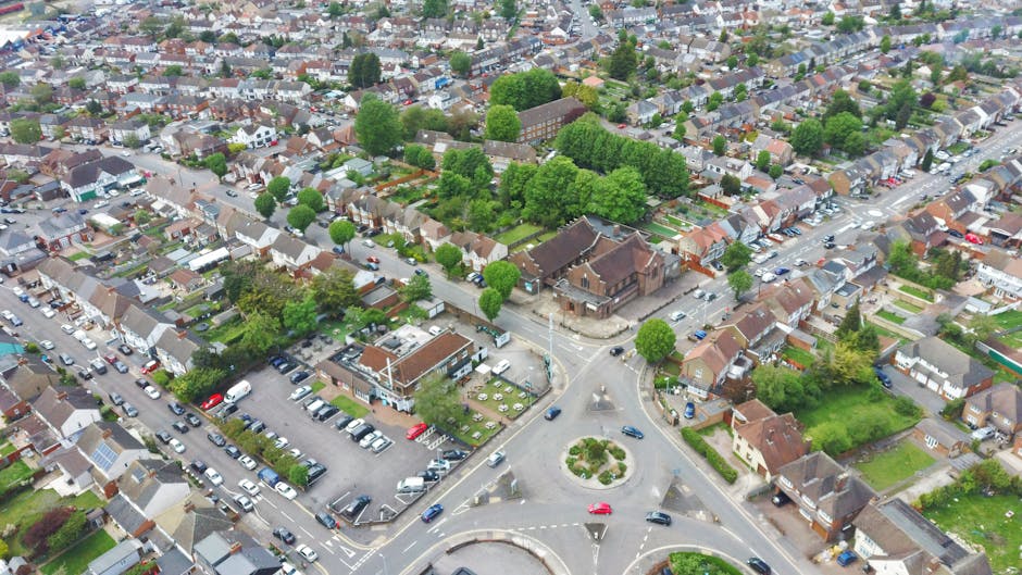 An aerial view of a residential neighbourhood in West Green showing a variety of houses, including terraced and semi-detached properties, with streets lined by trees and parked cars. In the foreground, a roundabout features a landscaped central island with bushes and grass, with several vehicles navigating around it. To the left, a multi-storey parking lot is filled with cars, adjacent to a small building likely used for community or commercial purposes. The scene captures the layout of the area during daylight, with natural lighting highlighting the rooftops, greenery, and paved roads. This image supports understanding of house layout, vehicle positioning, and the logistics involved in home relocations and furniture transport within West Green. It reflects the typical environment where professional removals, offered by Man with Van West Green, operate, especially regarding movement planning around local noise curfews and moving times.