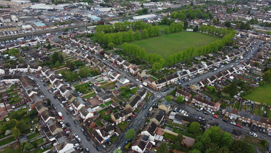 Aerial view of a residential neighbourhood in West Green featuring tightly packed terraced houses with small gardens, cars parked along narrow streets, and a large green park surrounded by a dense line of trees. The image shows the area’s urban layout with a mix of low-rise homes, side streets, and open recreational space, highlighting the typical environment for house removals and furniture transport. The scene captures the daytime with natural lighting, illustrating the typical setting where Man with Van West Green provides packing, loading, and house removal services, including careful handling of furniture and boxes during the home relocation process across this suburban area.