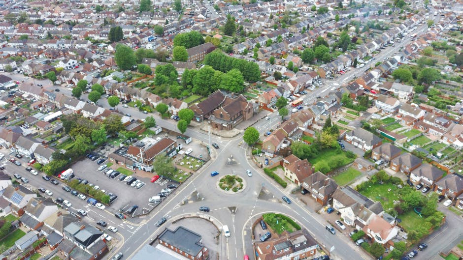 An aerial view of a residential neighbourhood in West Green showing a variety of houses, including terraced and semi-detached properties, with streets lined by trees and parked cars. In the foreground, a roundabout features a landscaped central island with bushes and grass, with several vehicles navigating around it. To the left, a multi-storey parking lot is filled with cars, adjacent to a small building likely used for community or commercial purposes. The scene captures the layout of the area during daylight, with natural lighting highlighting the rooftops, greenery, and paved roads. This image supports understanding of house layout, vehicle positioning, and the logistics involved in home relocations and furniture transport within West Green. It reflects the typical environment where professional removals, offered by Man with Van West Green, operate, especially regarding movement planning around local noise curfews and moving times.