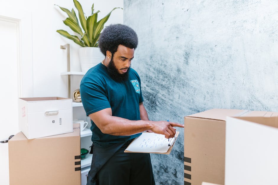 A man with dark, curly hair and a beard, wearing a navy blue t-shirt with a logo on the chest, stands inside a room surrounded by cardboard boxes and packing materials. He is holding a clipboard in hi