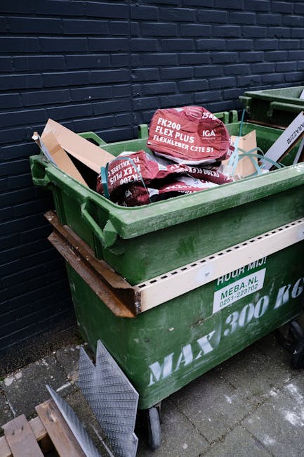 A large green wheelie bin positioned on a paved surface against a dark brick wall, filled with various items including cardboard boxes and 25-kilogram bags of fencing gravel with red and white packaging, some of which are slightly protruding from the bin. The bin is part of a house removal or waste disposal process managed by Man with Van West Green, with additional waste management containers visible nearby, and a piece of metal grating leaning against the side of the bin, indicating an outdoor setting suitable for loading and transport during home relocation or furniture transport activities.