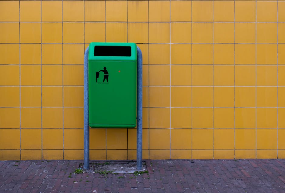 A green waste disposal bin with a black pictogram of a person disposing of rubbish is mounted on a yellow tiled wall. The bin is supported by two vertical metal posts anchored into the pavement, which is made of brick and shows some small patches of moss or dirt near the base. The bin has an open rectangular slot at the top for inserting waste and a small red indicator light on the front. This scene may be adjacent to a residential or commercial property, illustrating typical urban waste management. The context of house removals, such as packing or transport, can be associated with the setting where waste disposal is part of the moving or clearing process, which Man with Van West Green offers as part of their removals services.