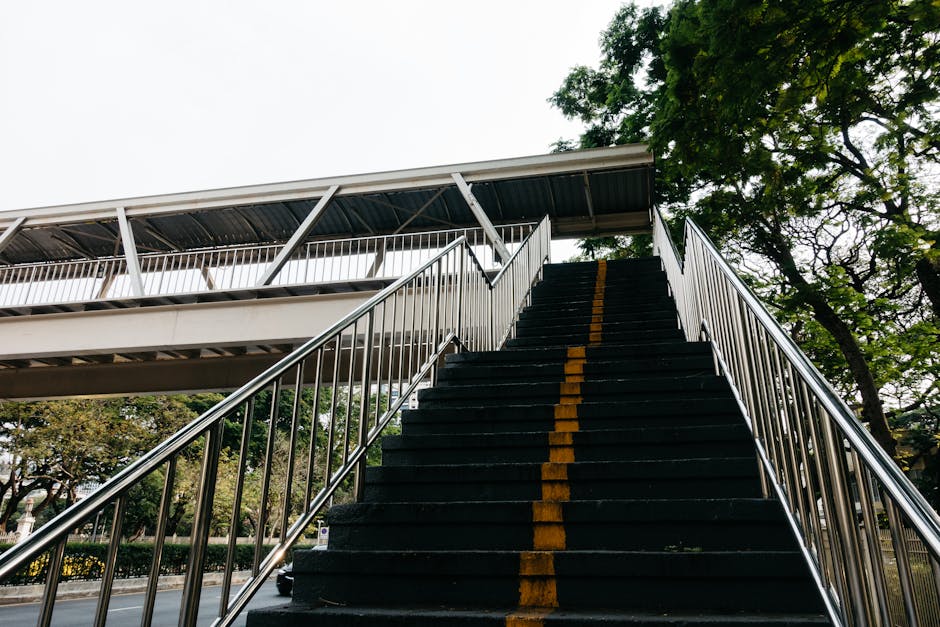 A staircase with light-colored wooden steps and a black metal handrail curves to the upper floor of a residential building, situated next to a window with natural light illuminating the area. On the l
